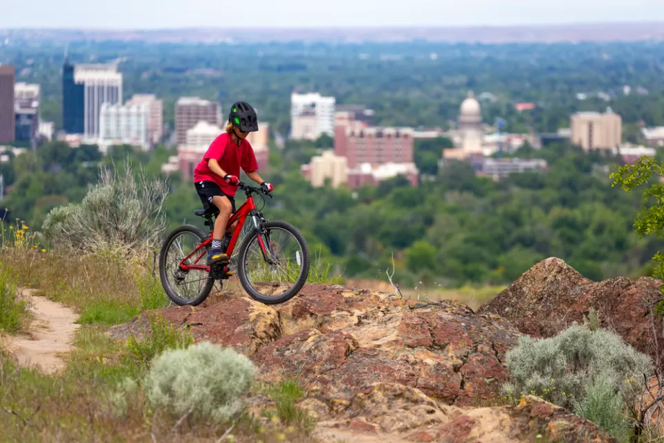 An adolescent boy catches some air on a mountain bike on a hill above the city of Boise. The city’s downtown is clearly visible in the background.